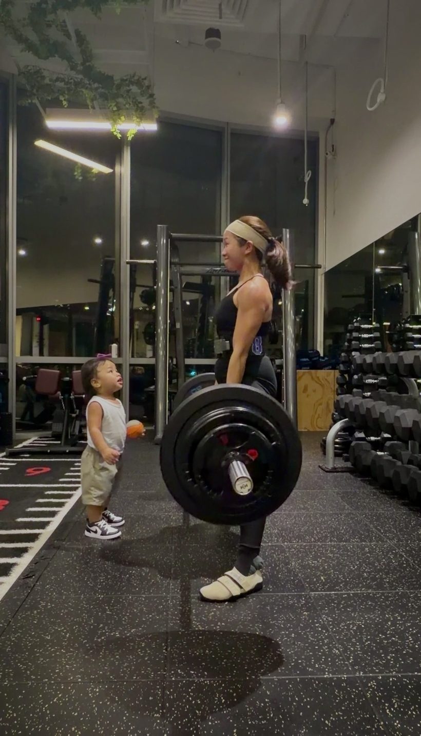 Patricia finishing a barbell deadlift while her toddler watches from the gym floor.
